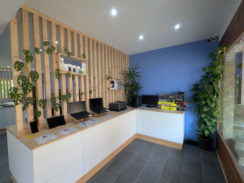 Ordinateurs portables et tablettes exposés sur un meuble blanc en L, devant un mur de tasseaux de bois et des plantes.